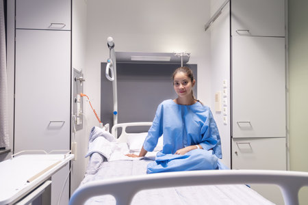 Young female patient in hospital ward,dressed in blue gown,sitting on bed and waiting to be examined by doctor,smiling softly and satisfied with service and services provided,individual hospital roomの写真素材