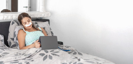 Woman resting in bed in casual clothes and bandages on her face after plastic surgery on her nose,using tablet to communicate with family, calm atmosphere and soft colors illustrate recovery processの写真素材