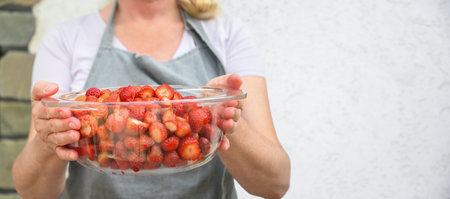 unrecognizable woman in an apron holds large transparent bowl of freshly picked strawberries, stemmed and ready to be made into jam,concept of new harvest of seasonal berries and organic gardeningの写真素材