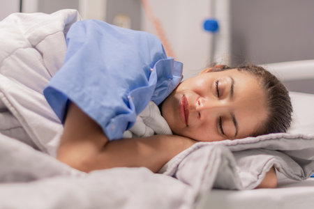 Young woman in blue robe lying comfortably and getting ready for bed on hospital bed in calm environment of clinic in private room, soft lighting, concept of recovery and care,の写真素材