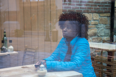 An African American woman in a blue jacket sits behind a window glass in a cafe waiting for a...