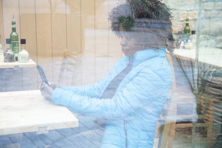 An African American woman in a blue jacket sits behind a window glass in a cafe makes an order using an application on a smartphone, selective soft focus from due to dirty glass. High quality photoの写真素材