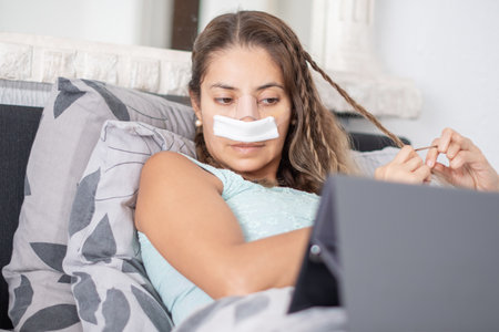 woman lies in bed in casual clothes and with bandages on her face, braids hair and watches TV on tablet,の写真素材