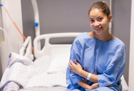 young beautiful woman in private hospital room, dressed in blue robe, sits on bed and rejoices at quick correction of cosmetic defect, waits for doctors examination before plastic surgery,の写真素材