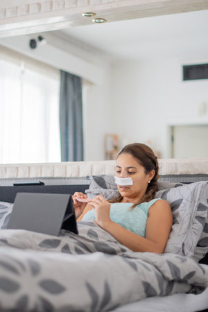 woman lies in bed in casual clothes and with bandages on her face, files nails and watches favorite movie on tablet,の写真素材