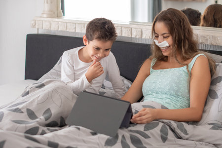 Smiling mother with nasal strips on her face after rhinoplasty lies in bed with her little son and watches movie on digital tablet, concept of joint leisureの写真素材