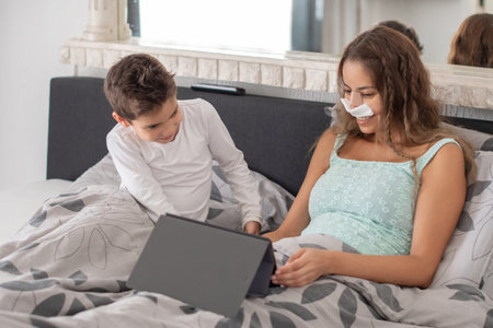 Smiling mother with nasal packing on her face lies in bed with her little son and watches movie on digital tablet, concept of joint leisureの写真素材