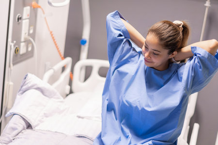 Young woman in blue gown on hospital bed tying it behind her back smiling cheerfully preparing to be examined by doctorの写真素材