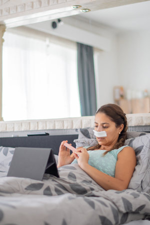 woman lies in bed in casual clothes and with bandages on her face, files nails and watches favorite...