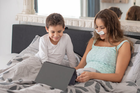 Smiling mother with bandage on nose lying in cozy bed and helping her schoolboy son to do homework, concept of upbringing and education, family relaxing lying among pillows,の写真素材