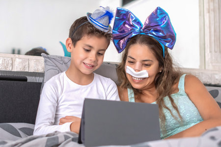 Smiling mother with bandage on nose lies in cozy bed and plays on tablet with son, mother has big blue bow on head and son has blue hat, carnival in bed to lift mood, relaxing,の写真素材