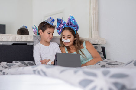 Smiling mother with bandage on nose lies in cozy bed and plays on tablet with son, mother has big blue bow on head and son has blue hat, carnival in bed to lift mood, relaxing,の写真素材