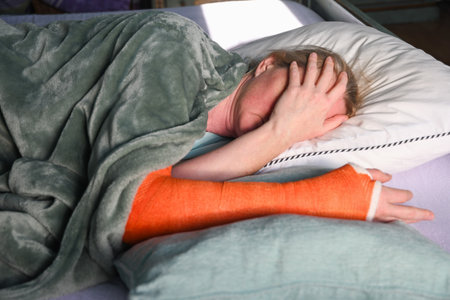 woman with a broken right arm with an orange fiberglass plaster cast sleeps in bed with a pillow under the cast, everyday life with trauma,の写真素材
