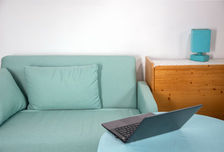 Modern interior with turquoise sofa, laptop on table, wooden cabinet and turquoise lamp on white background, home workspace concept, remote work from homeの写真素材