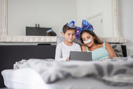 Smiling mother with bandage on her nose lies in cozy bed and watches TV on tablet, mother has big blue bow on her head and son has blue hat, carnival in bed to lift mood, relax,の写真素材
