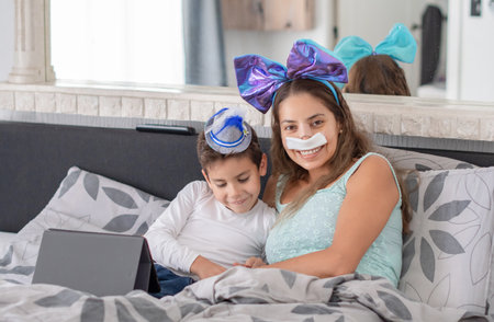 Smiling mother with bandage on her nose lies in cozy bed and watches TV on tablet, mother has big blue bow on her head and son has blue hat, carnival in bed to lift mood, relax,の写真素材