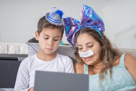 Smiling mother with bandage on her face after rhinoplasty lies in bed with her little son and watches movie on digital tablet, concept of joint leisureの写真素材