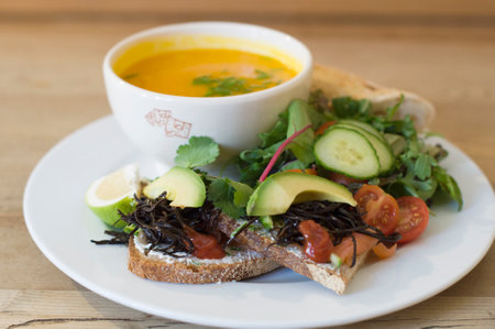 Yellow pumpkin soup in a ceramic bowl on a plate with a fresh vegetable salad of avocado, tomatoes, cucumbers and arugula, as well as pieces of toasted white bread spread with butter,の写真素材