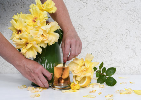 hand stirs lemon tea in a glass, bouquet of yellow roses stands in a green vase next to a glass of tea with lemon, scattered rose petals, creating a harmonious still life on a white table,の写真素材