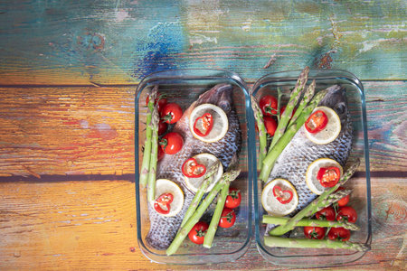 Green asparagus on fresh large dorado fish, tilapia, carp in baking dish with cherry tomatoes,step by step recipe instructions, top view on an old wooden tableの写真素材