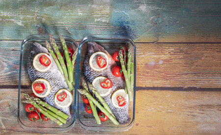 Raw green asparagus on fresh large dorado fish, tilapia, carp in a baking dish with cherry tomatoes, top view on an old wooden tableの写真素材