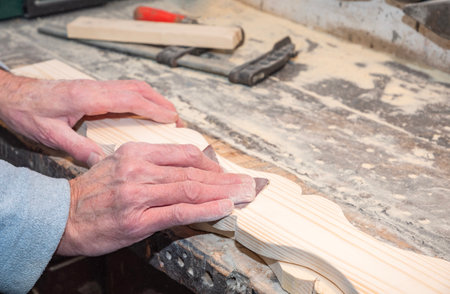 Close-up of bare carpenters hands skillfully sanding decorative wooden board on workbench in carpentry workshop, surrounded by various tools and small sawdustの写真素材
