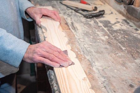 Close-up of carpenters hands skillfully sanding decorative wooden plank on workbench in carpentry workshop, surrounded by various tools and small sawdustの写真素材