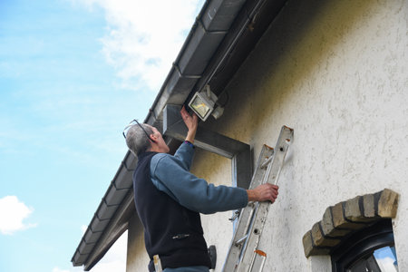 Electrician checks the settings of a motion sensor LED floodlight while standing on a ladder, the...