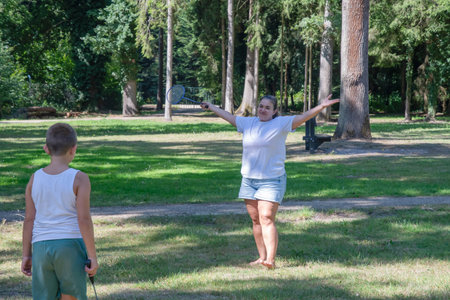 Mother and son playing badminton barefoot on green grass in a sunny park, laughing and bonding...