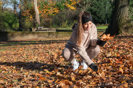 Young woman in a tracksuit and headband crouching on the ground in a public park, picking up and holding a large fallen maple leaf during the vibrant autumn seasonの写真素材