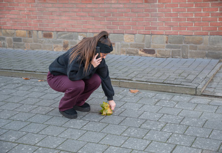 Blindfolded woman crouching on the paved sidewalk, engaged in a phone conversation while attempting to pick up autumn leaves, representing the challenges of multitasking and modern distractionsの写真素材