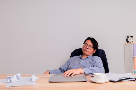 Portrait of a tired Asian businessman sitting at his desk and using laptopの写真素材