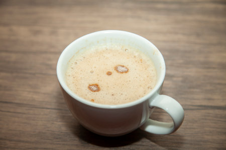 Frothy cappuccino with some powder not mixed in well in a white cup on a wooden background