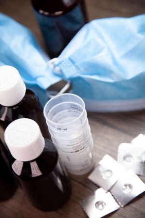 Cough medicine, medication cups, and open and empty medication blister packs with a mask in the backgroundの写真素材