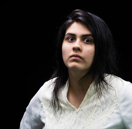 Latina teenager in white standing in front of a black background with an angry, suspicious, serious...