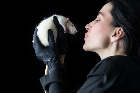 Brunette woman in black standing in front of a black backdrop, holding a white dumbo rat, looking at it with affection and love.の写真素材