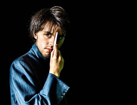 Low key shot of a young man standing in front of a black backdrop, holding a card up to his eye and looking at the cameraの写真素材