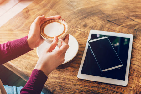 Woman hand holding cup coffee in cafe with smartphone and tablet on wood table.