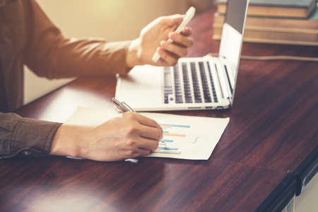 Young business man hand using phone and writing paper in the office with sunlight. Vintage toned filter.の写真素材