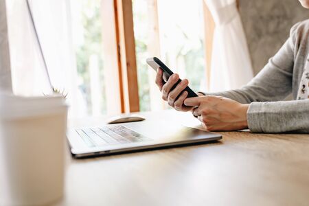 Asian woman holding phone and using computer laptop on wooden tableの写真素材