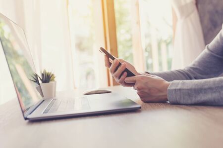 Asian woman holding phone and using computer laptop on wooden tableの写真素材