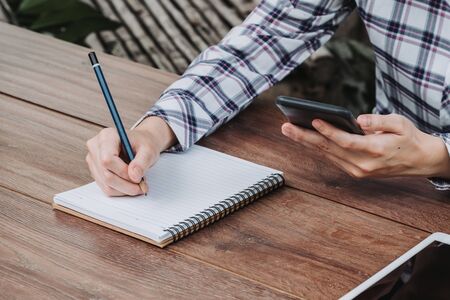 woman hand writing notepad and holding phone on wooden table at coffee shopの写真素材