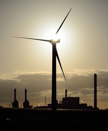 Wind turbine in foreground and power plant in background at sunriseの写真素材