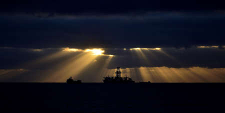Sunrise seen from the coast with beams of light between the clouds and backlit shipsの写真素材