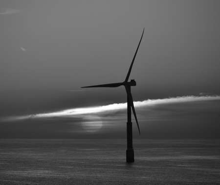 Backlit offshore turbine and sun behind the clouds at dawn, black and white modeの写真素材