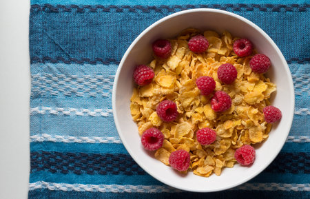 Breakfast cereal of cornflakes and raspberries on blue table cloth background
