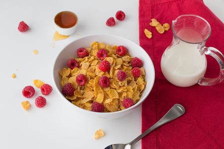Breakfast cereal of cornflakes, raspberries, honey and milk on white backgroundの写真素材