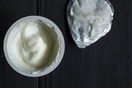 Yogurt cup with natural Greek yoghurt in plastic cup with foil lid closeup on rustic black table background - Top view photoの写真素材