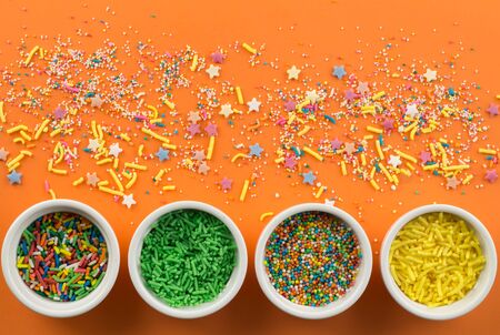 Sprinkles on bright orange background with little white bowls with various type of coloured sprinkles at bottom  and multi-colored cake topping sprinkles and star shapes sprayed aroundの写真素材