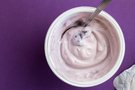 Yogurt cup with blue berry yoghurt, spoon and foil lid isolated on purple background with text space...